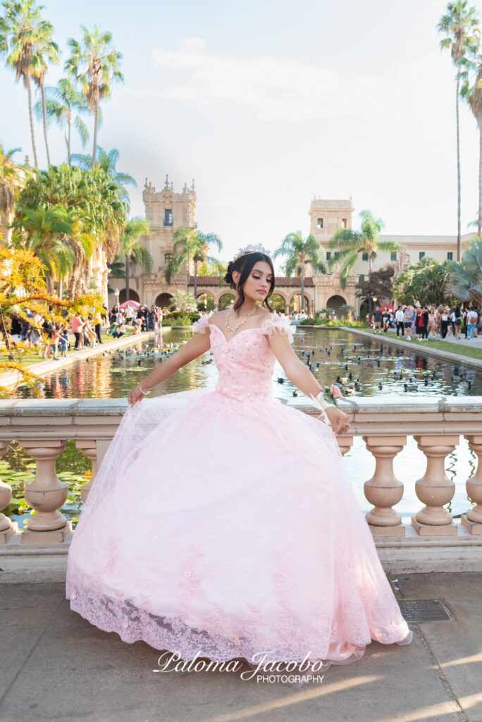 Picture of a Quinceanera wearing an pink dress with the lily pond on the background in a busy day at Balboa Park