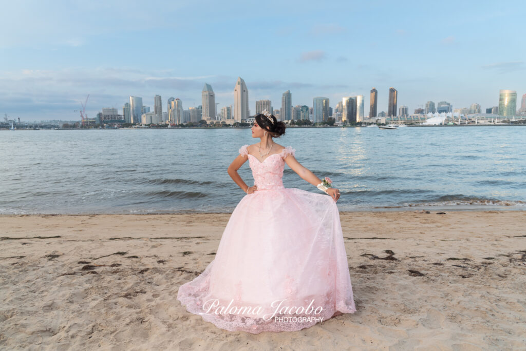 Quinceanera wearing an elegant pink dress posing on a sandy area with the San Diego city on the background